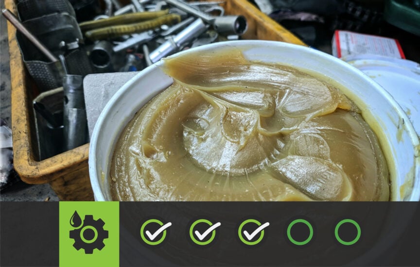 A photo of a five-gallon pail of grease. The grease is sitting on the floor of a dirty shop with a bin of tools behind it.